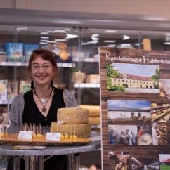 Eine lächelnde Frau steht an einem Tisch mit Bio-Käseprodukten, dahinter ein Aufsteller vom Kanzleilehngut Halsbrücke